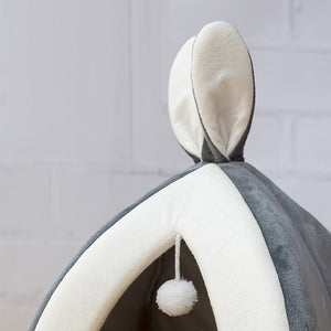 Gray and white pet bed with a pom-pom hanging from it against a gray background
